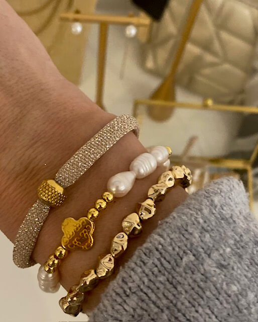 Eine Hand trägt mehrere elegante Armbänder, die mit goldenen und perlenbesetzten Details im Kosmetikstudio glänzen.